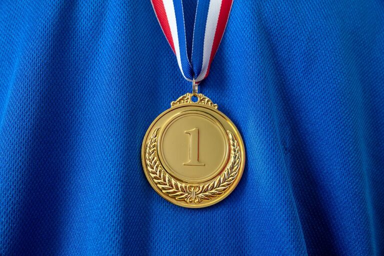 a gold 1st medal pictured on a blue textured fabric background, with a red white and blue ribbon
